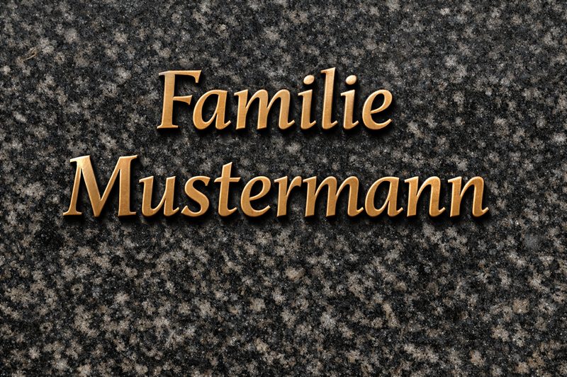Bronzeschriften auf einem Granitgrabstein: erhabene Buchstaben 'Familie Mustermann' mit warmer Patina, als Beispiel für Metallschriften